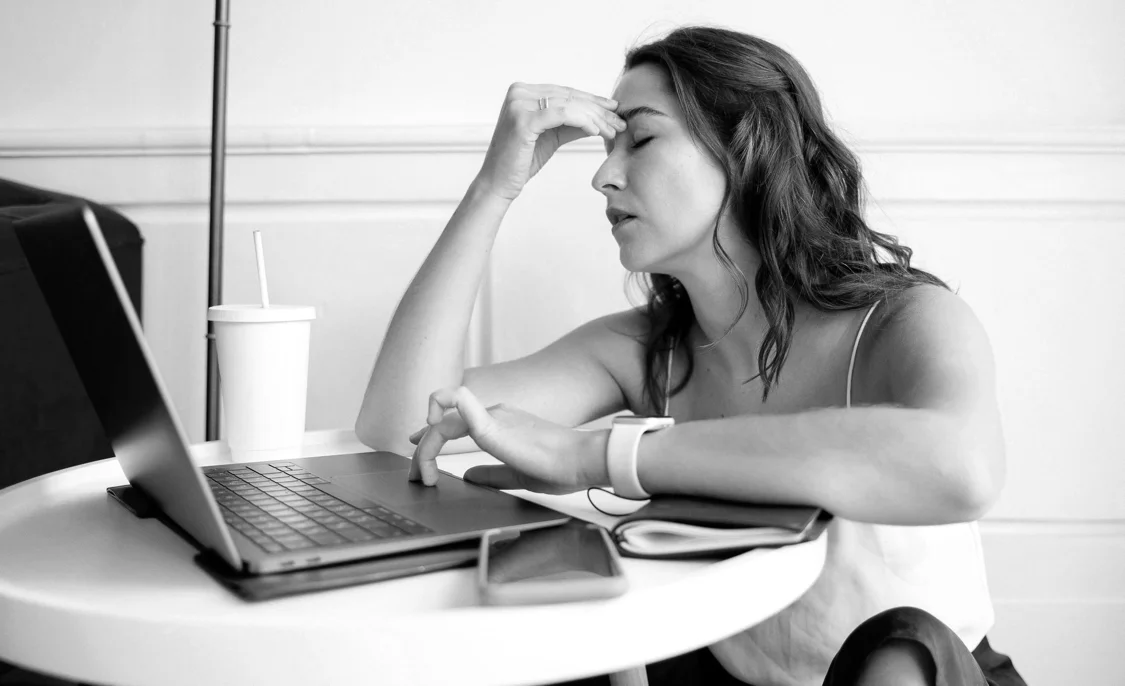 A woman with her hand on her head and her eyes closed, sitting at her laptop and looking stressed.