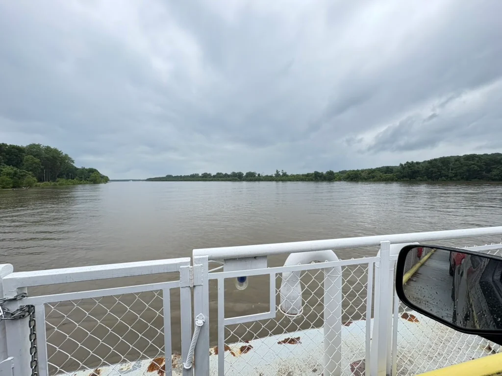 A wide, rain-grey river under heavy clouds, seen from the deck of the Brussels Ferry, with the ferry's white railing and a truck's wet side mirror in the foreground.