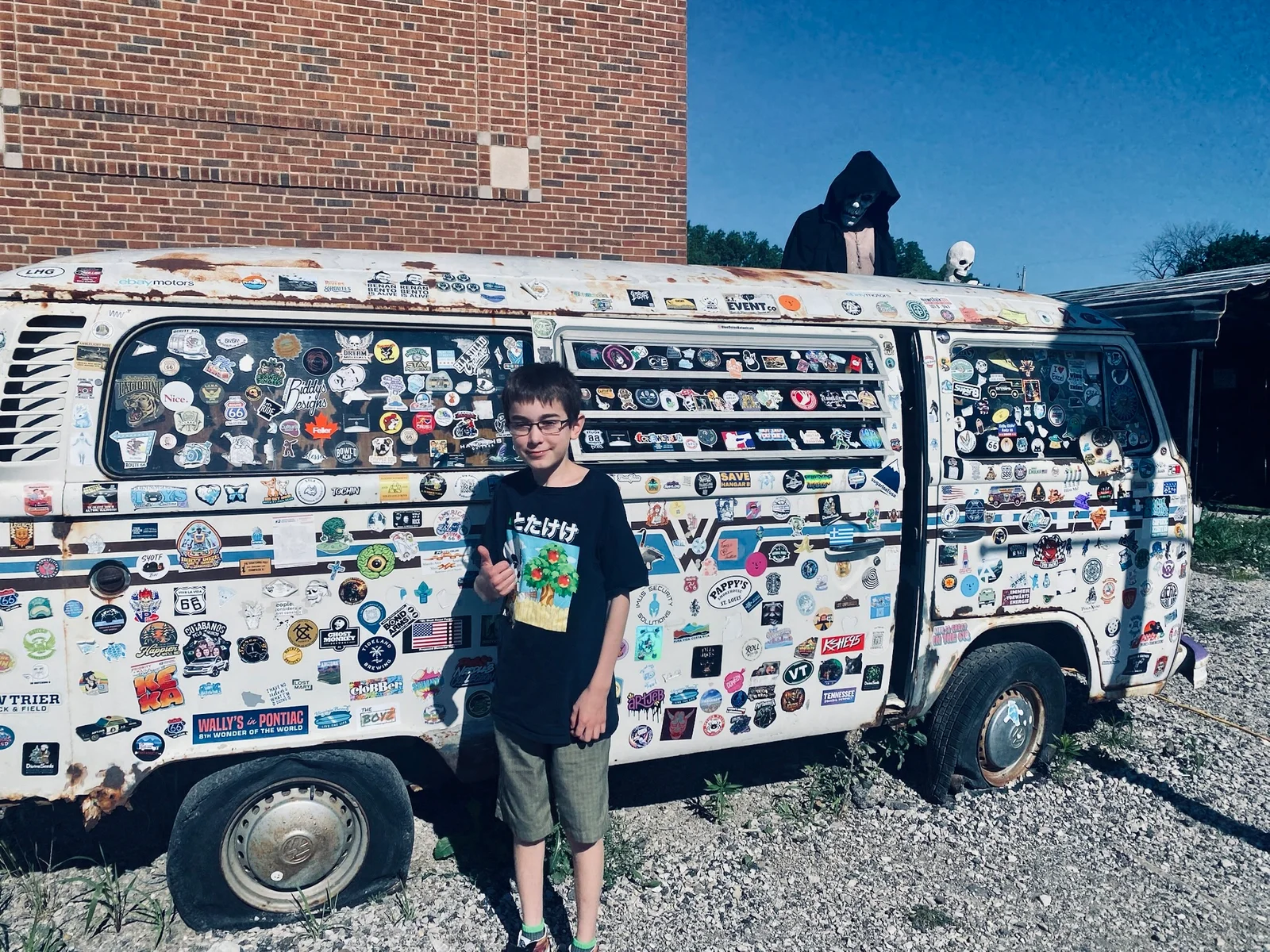 My son giving a thumbs up in front of an old VW bus covered roof to wheels in hundreds of stickers.