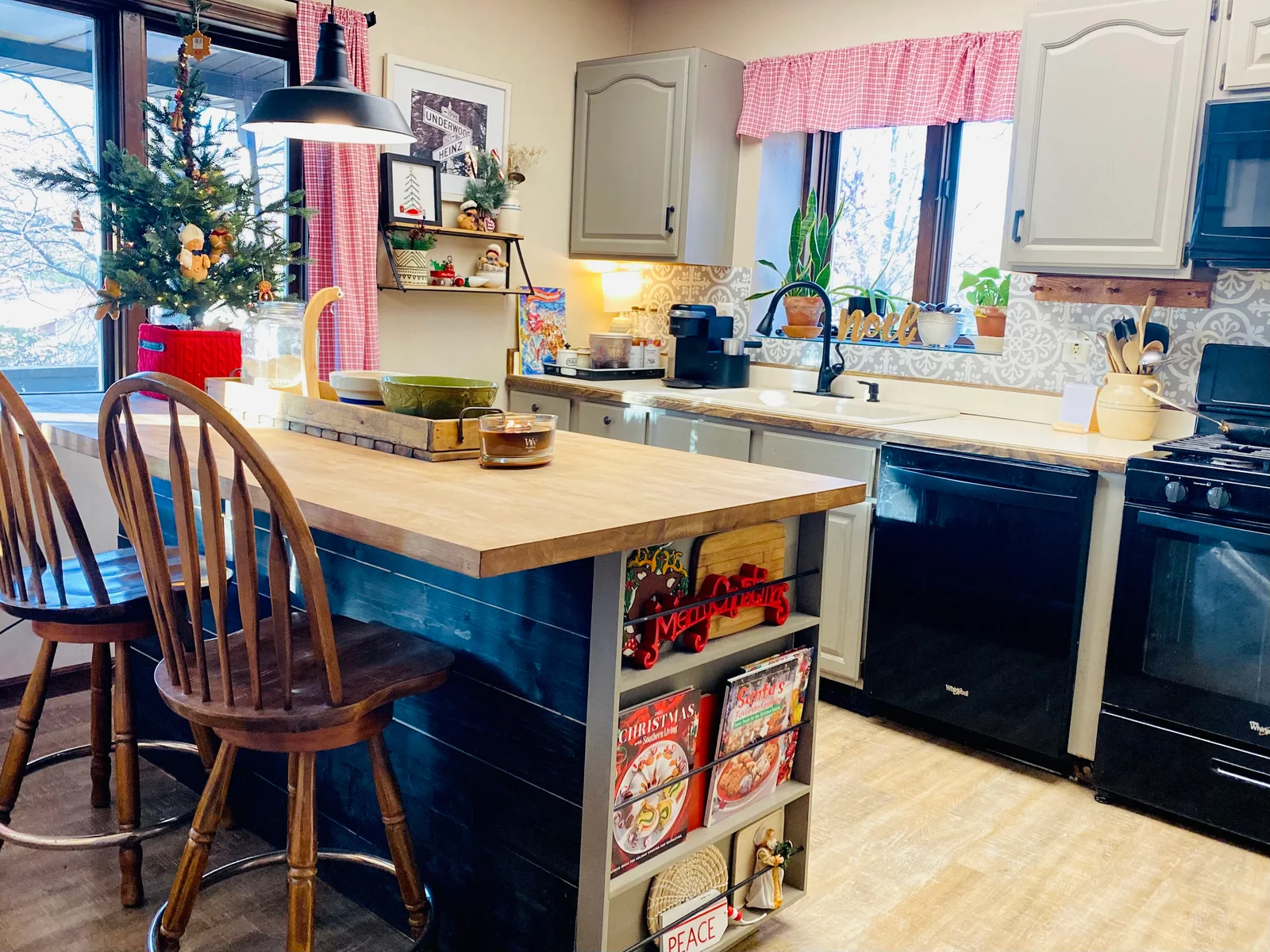 Warm, well-lit photo of a cozy kitchen featuring a large wooden island with two wooden bar stools in the foreground. A small decorated Christmas tree sits near the window on the left, with red gingham curtains, festive signs including “Merry Christmas” and “PEACE,” and various holiday decorations throughout the space.