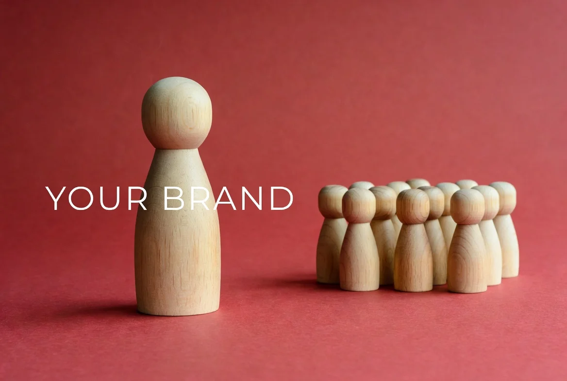 A larger wooden peg figure labeled ‘YOUR BRAND’ stands on the left, leading, while a cluster of smaller identical peg figures stands together on the right against a solid red background.