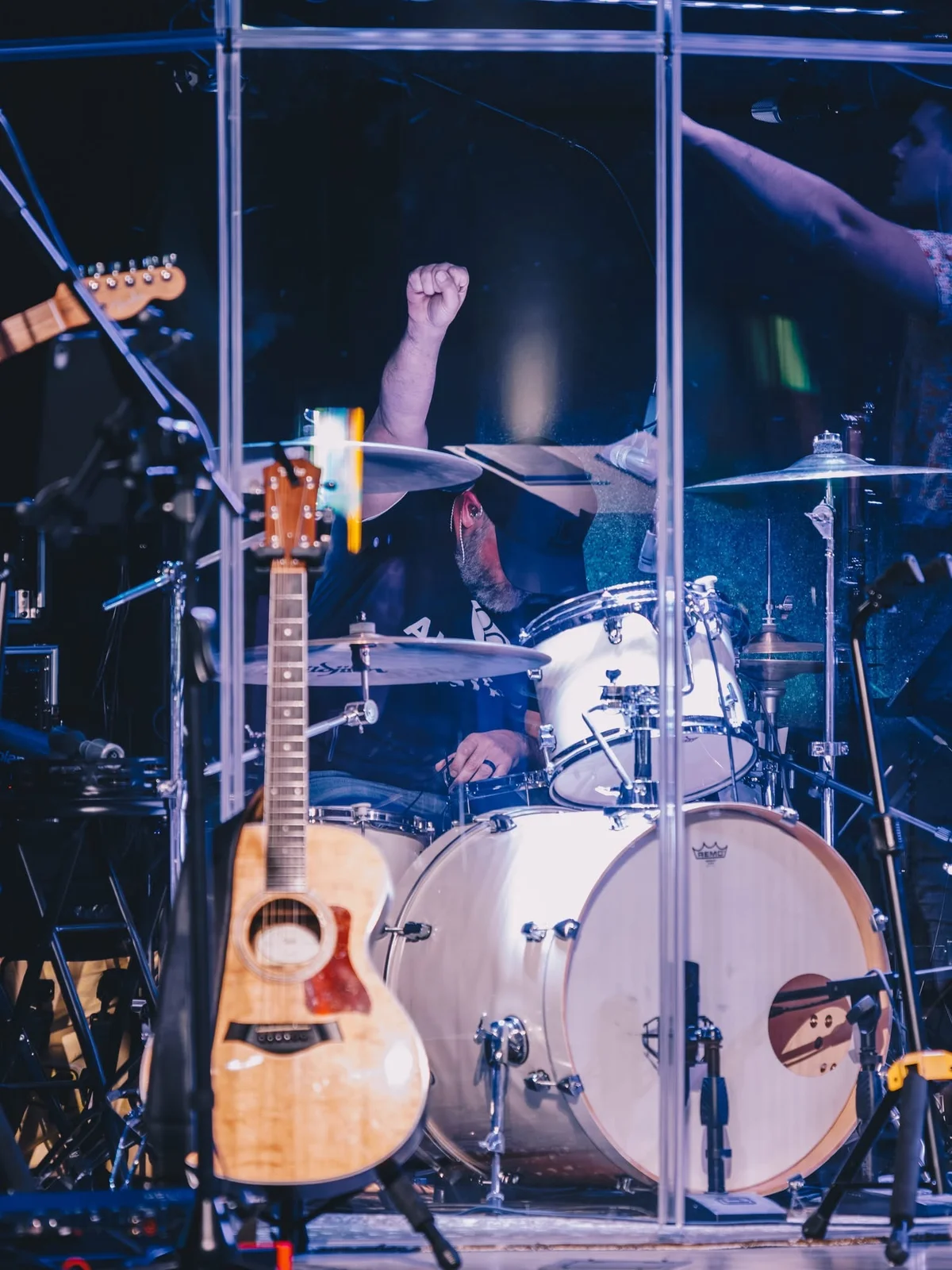 Drummer performing in a glass-enclosed booth on stage, with an acoustic guitar visible to the left and drum kit in foreground.