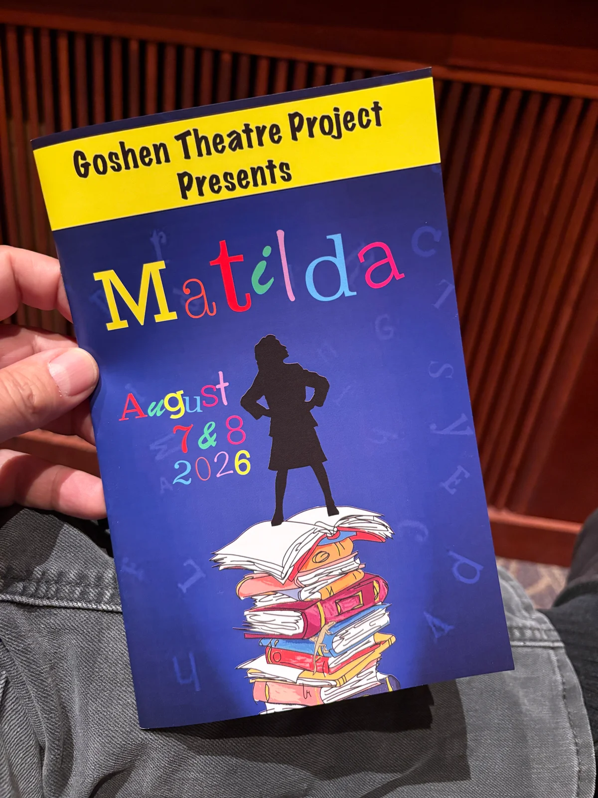 Program for Goshen Theatre Project's production of Matilda, scheduled for August 7 & 8, 2026, showing a silhouette of a girl standing on a stack of colorful books.