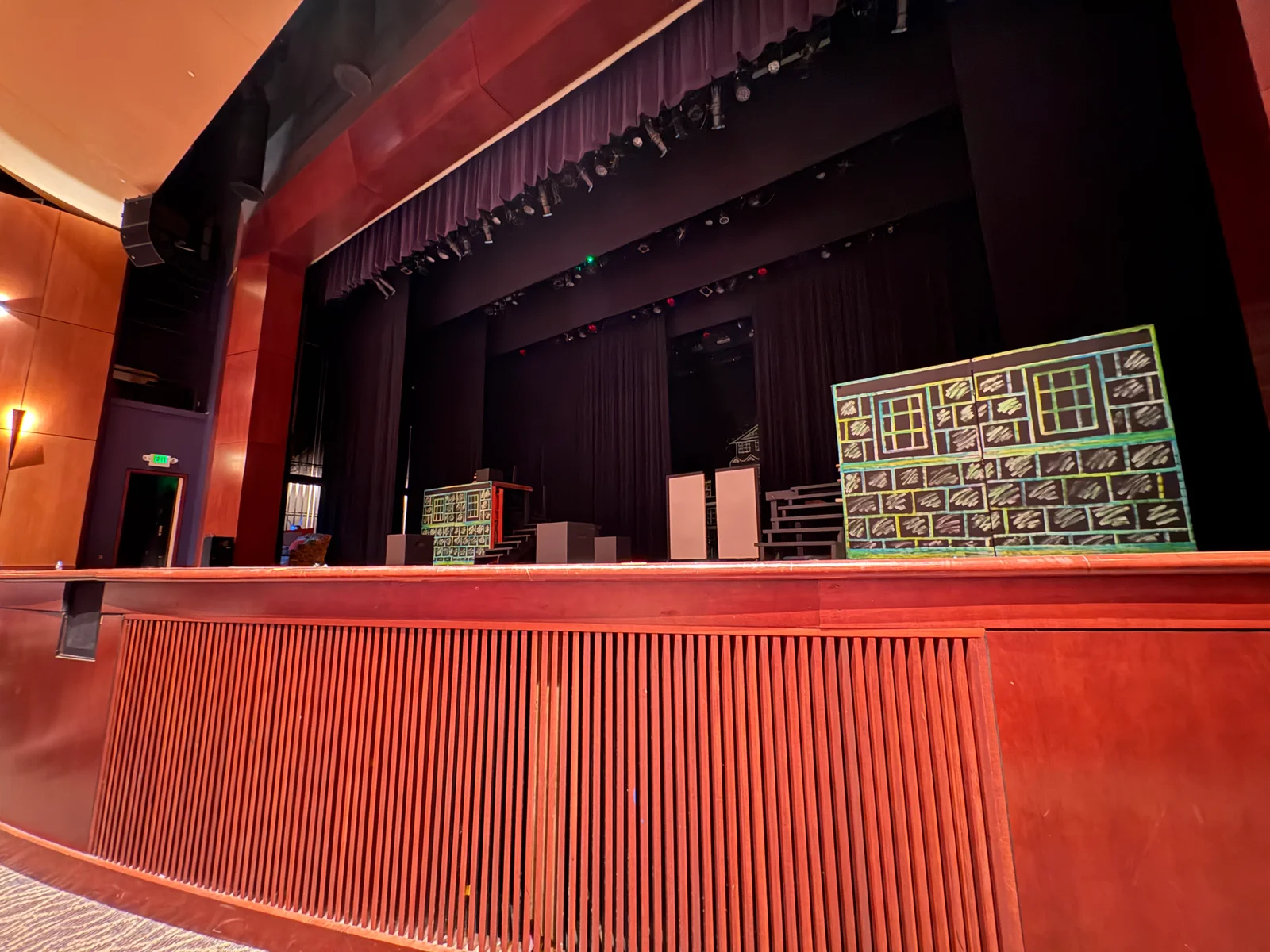 Theater stage with red barrier in foreground, purple curtains, hanging lights, and a large yellow-framed grid display on the right side.