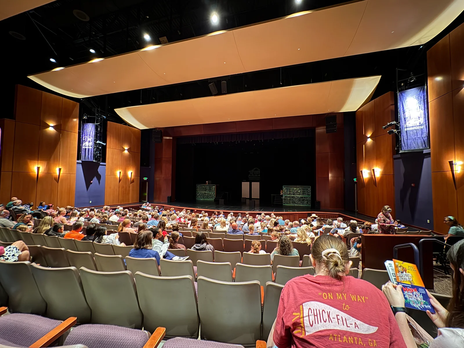 Audience seated in a theater auditorium with a black stage curtain and lit ceiling panels visible above.