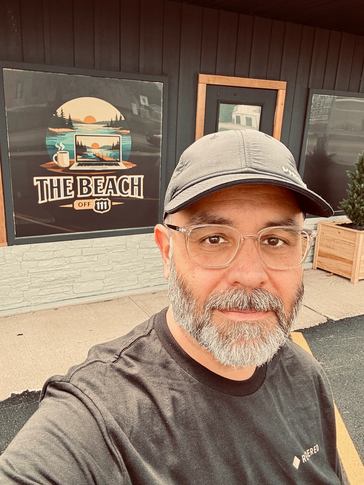 A photo of me wearing a black cap taking a selfie in front of a storefront with a "The Beach Off 111" sign featuring a sunset and laptop illustration.