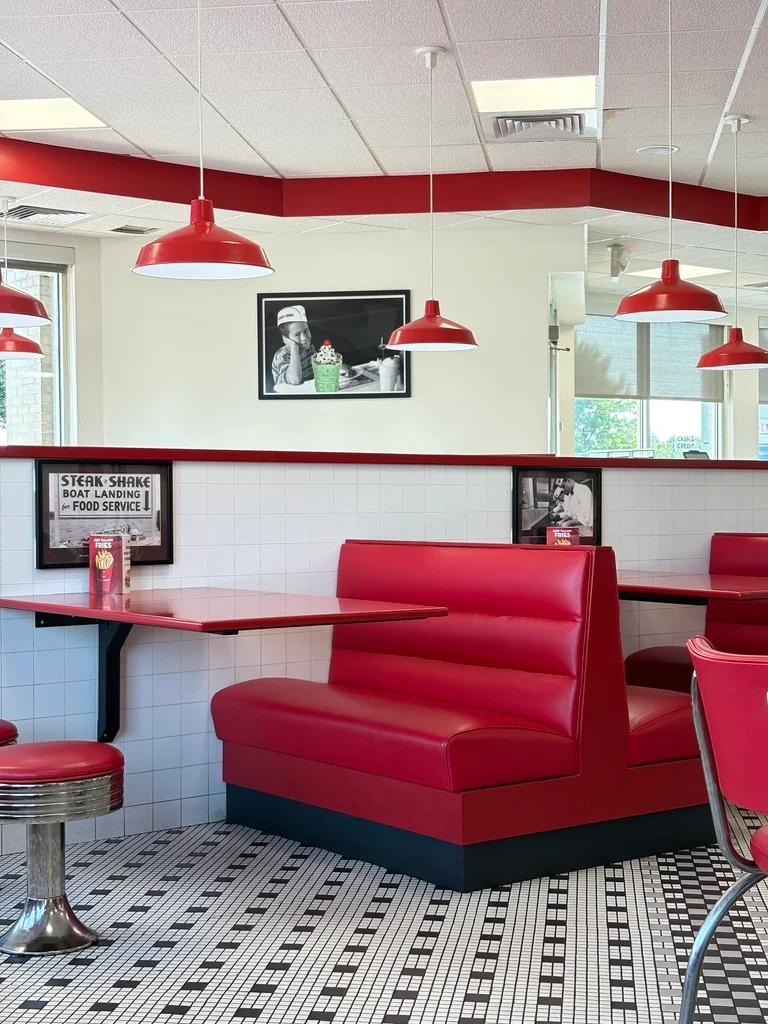 Retro diner interior with red vinyl booths, red pendant lamps, black-and-white checkered floor, and vintage signage on white subway tile walls.