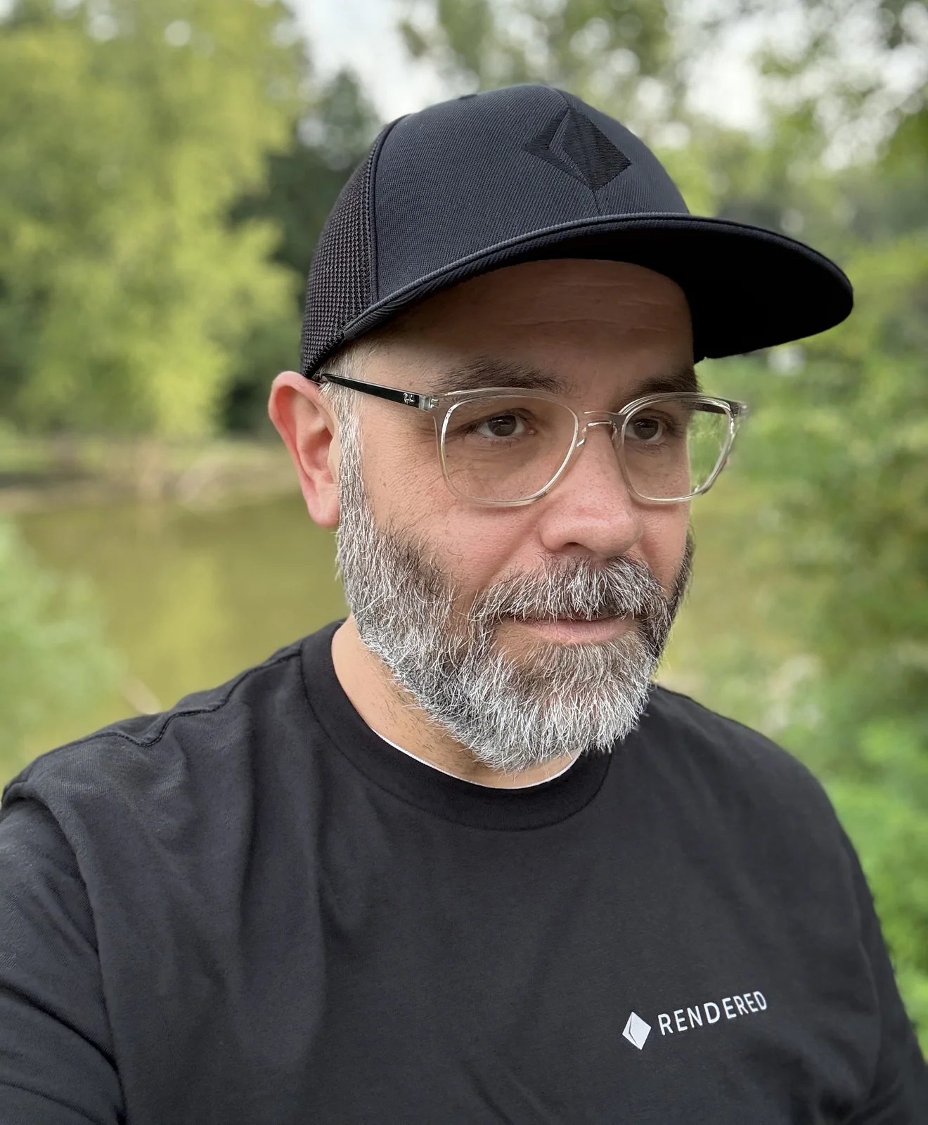 Me in the new Rendered gear: a black cap with the prism stitched tone on tone, and a black tee with the small Rendered lockup on the chest, outdoors in front of blurred trees and a pond.