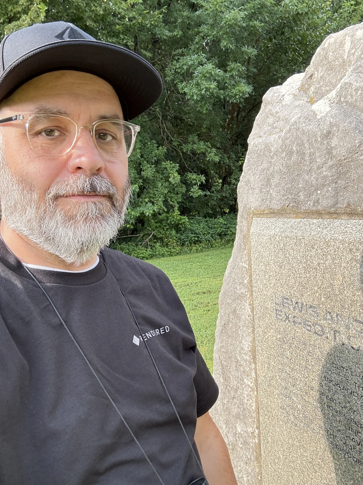 Me beside the stone Lewis and Clark Expedition marker at the site, its carved inscription weathered and faint, wearing the black Rendered cap and tee.