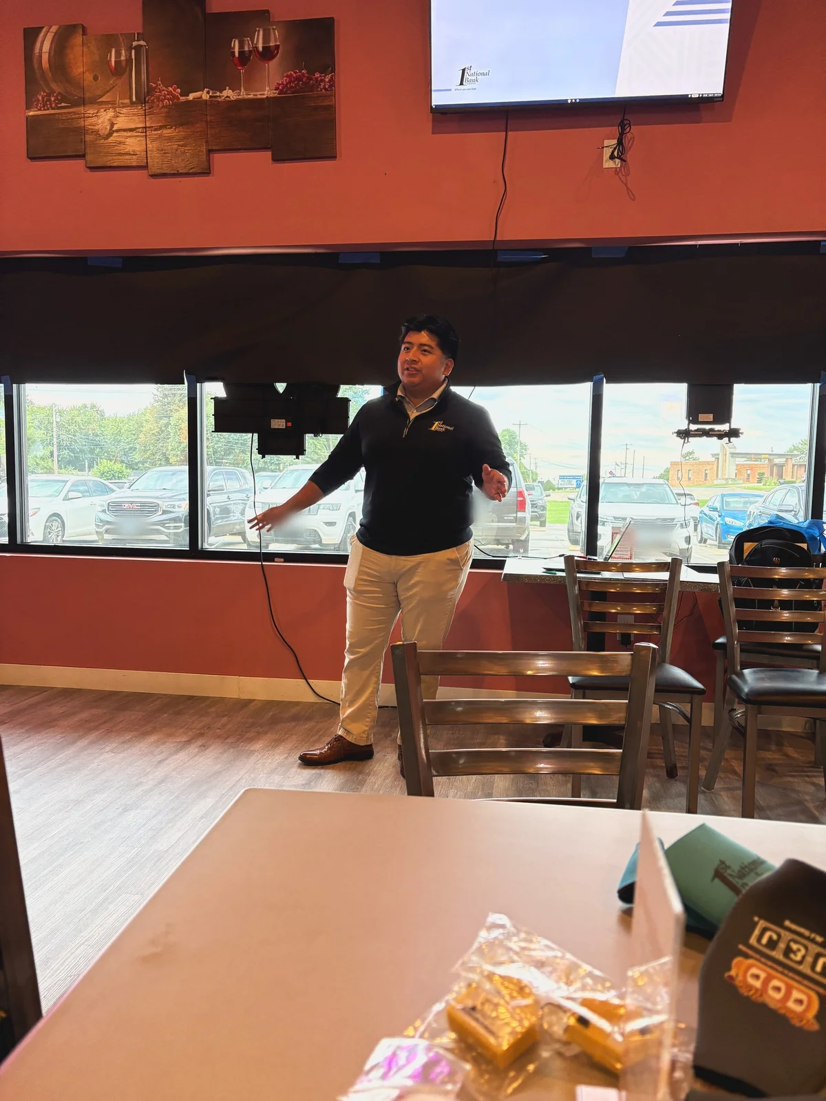 Marino Rojas stands and gestures while speaking to a room of chamber members at Wine for the Win in Troy, with a First National Bank of Waterloo slide on a TV behind him.