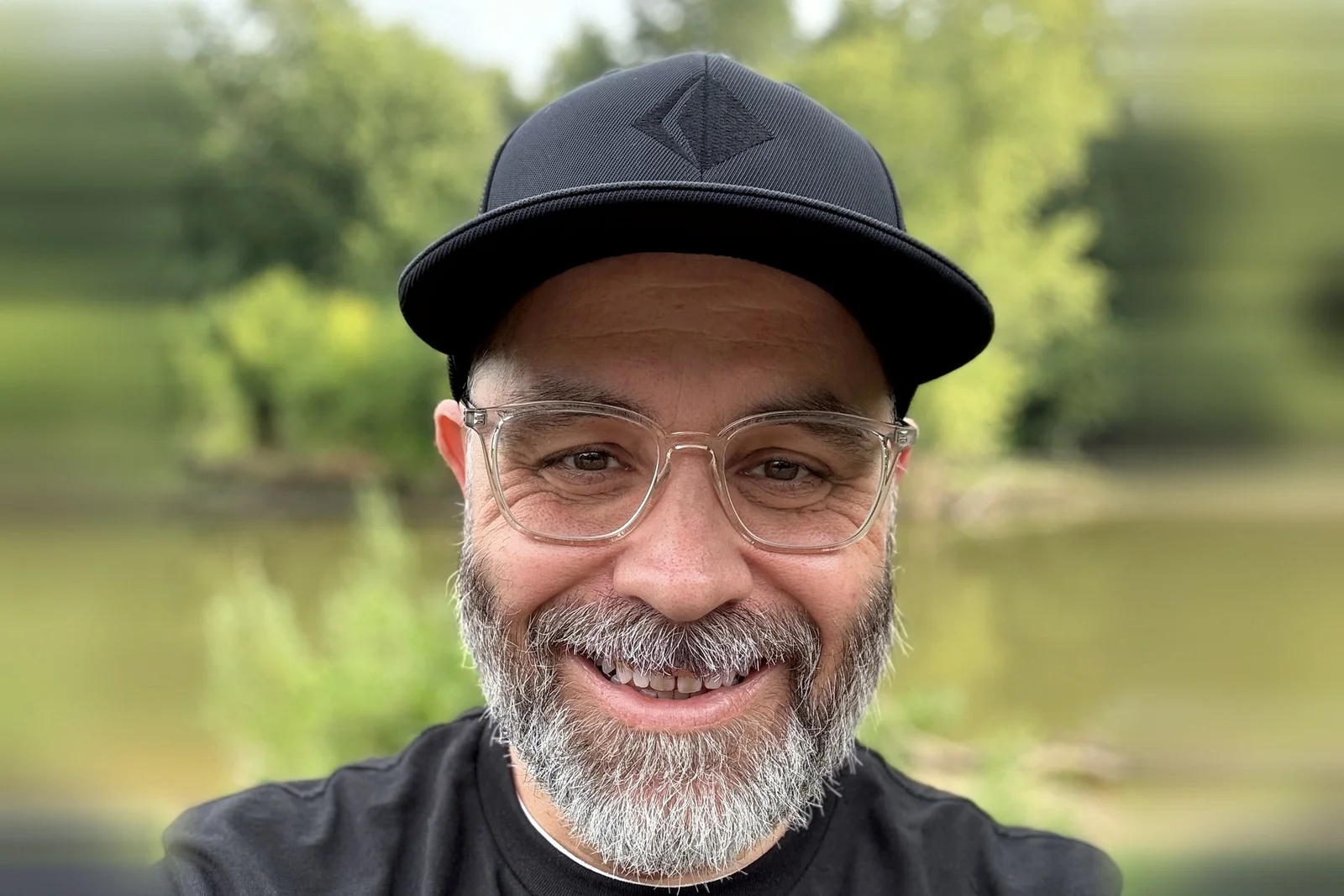 Mark Heinz smiling in a black Rendered cap and clear-framed glasses, a river and green trees soft-focus behind him.