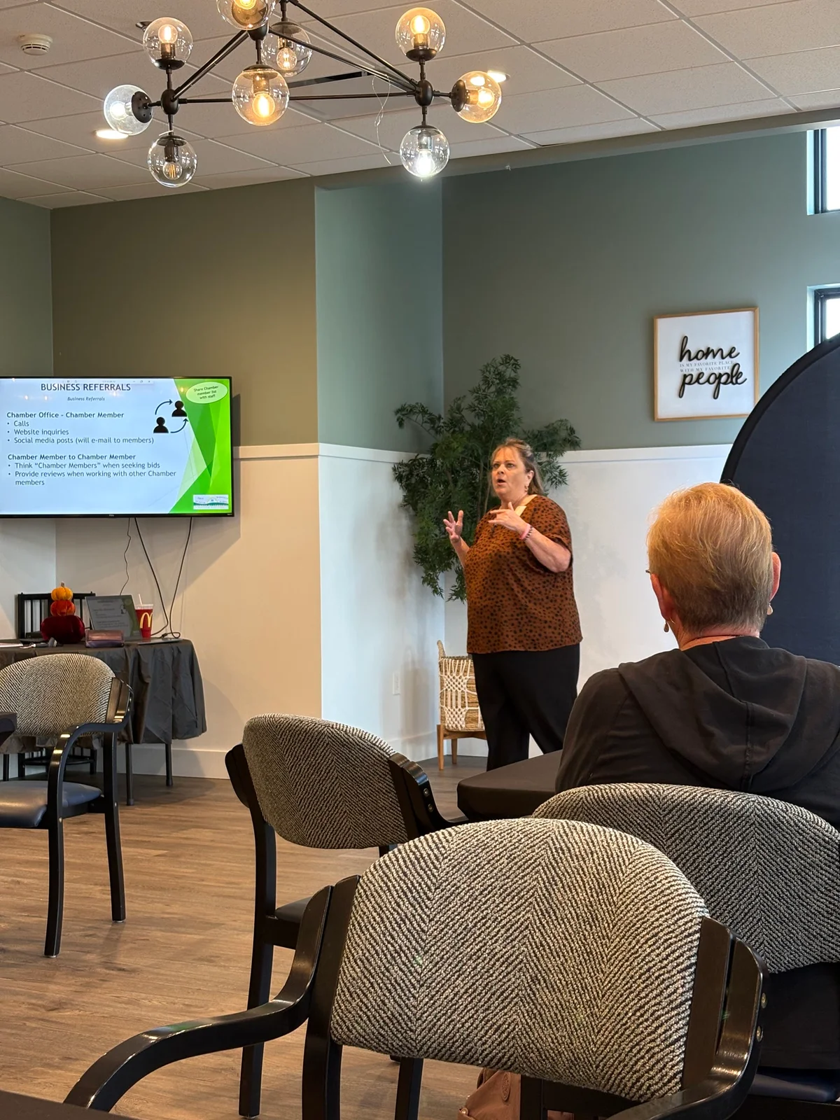 Dawn Mushill speaks to new members beneath a globe chandelier, with a Business Referrals slide on a TV nearby.