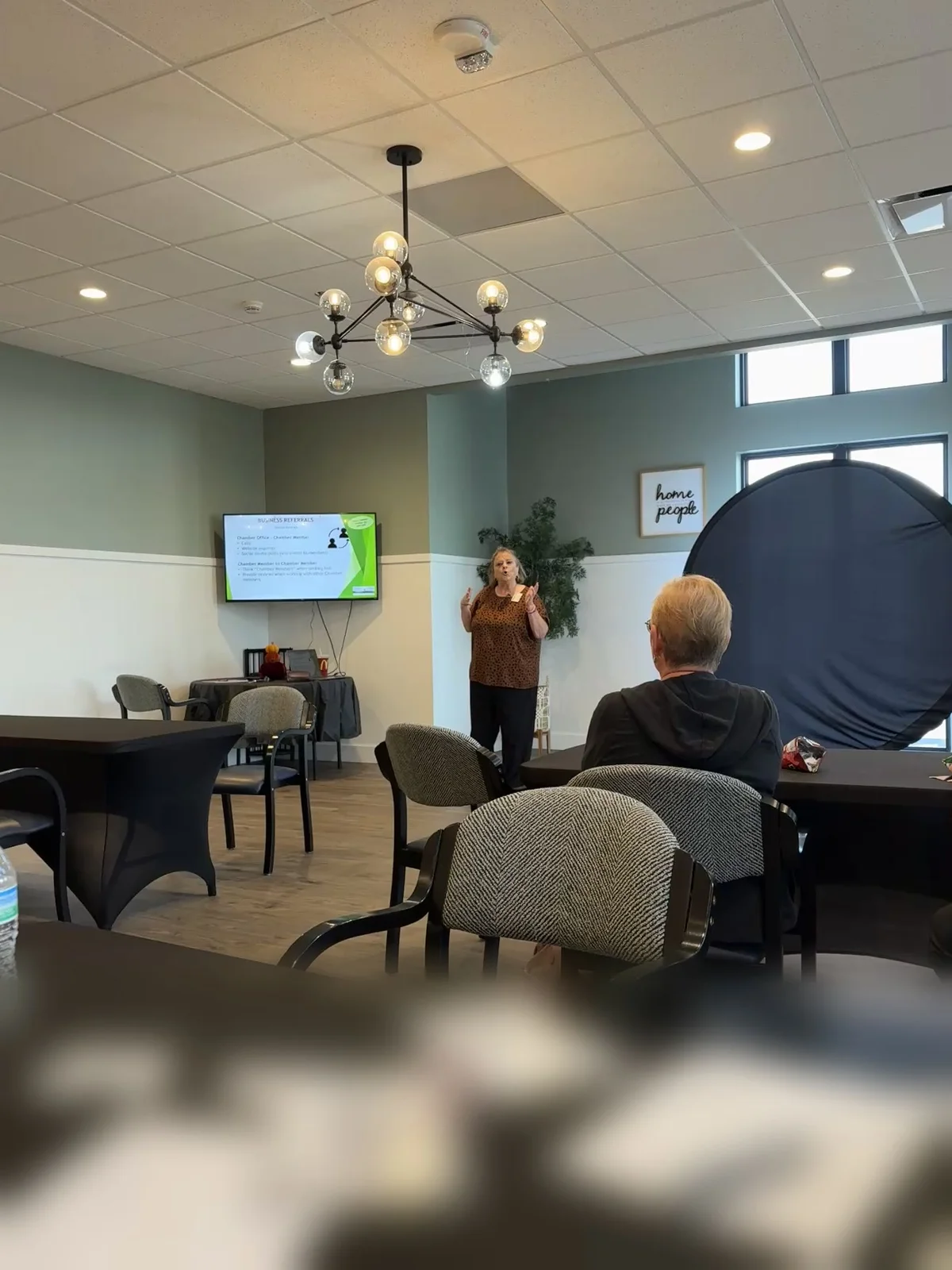 The view from a seat at a black-draped table toward the presenter at the front of the room, with the table in the foreground out of focus.