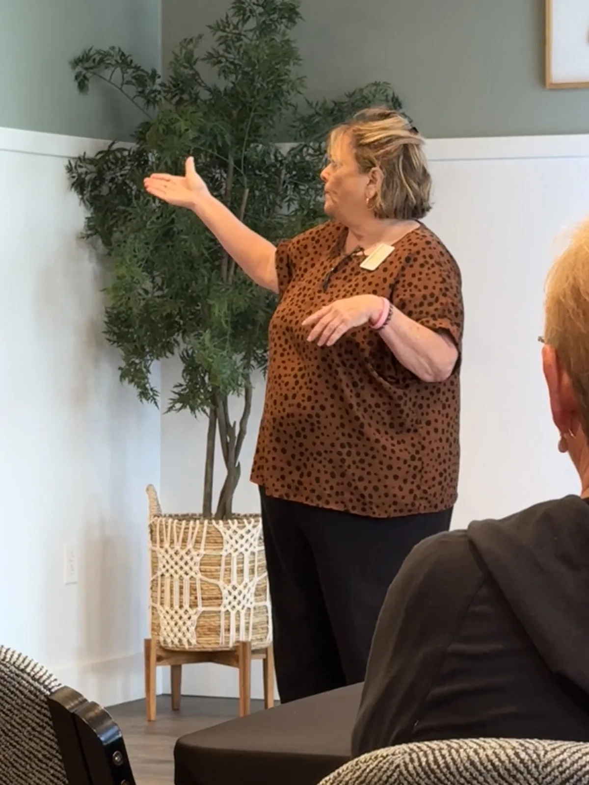Dawn Mushill gestures toward the room while speaking, in front of a potted tree in a macrame basket.