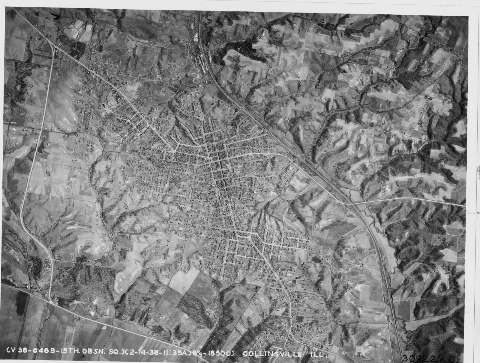Black-and-white aerial photograph: the street grid of a small town in the middle of farm fields and wooded ravines, with one long straight road cutting across the right side.