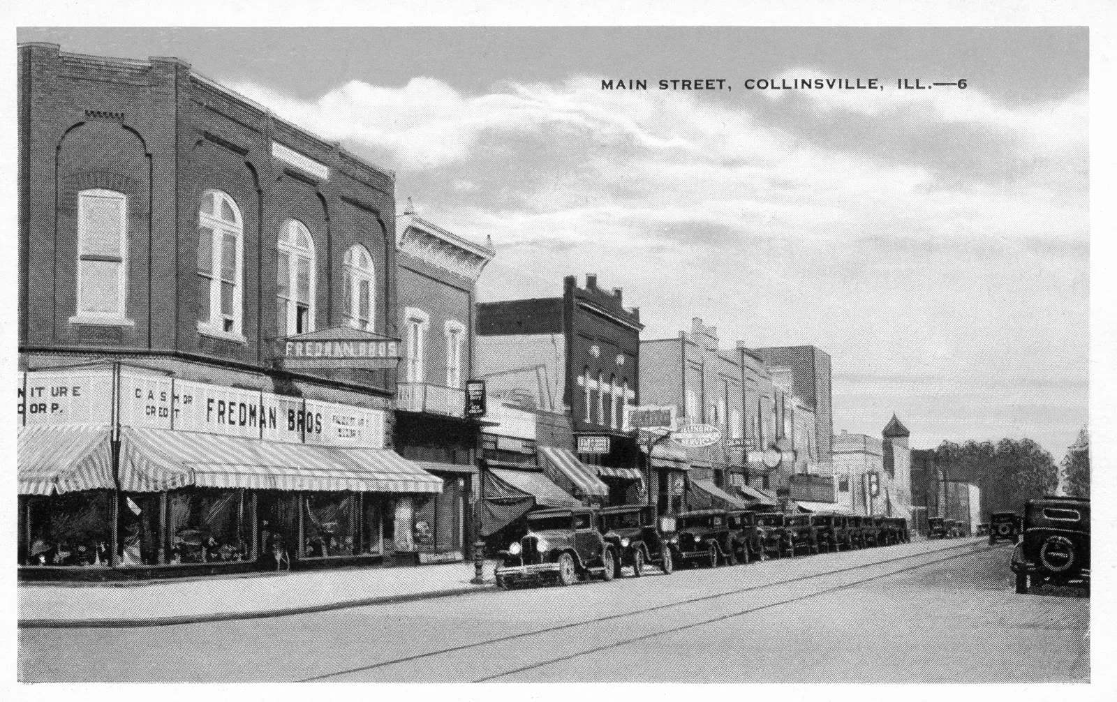 Black-and-white postcard view down a small-town Main Street: two-story brick storefronts with striped awnings, Fredman Bros. furniture store at left, a line of 1920s automobiles at the curb, and streetcar tracks in the pavement.